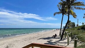Una playa con un paseo de madera que conduce a la orilla, palmeras y un cielo azul despejado.