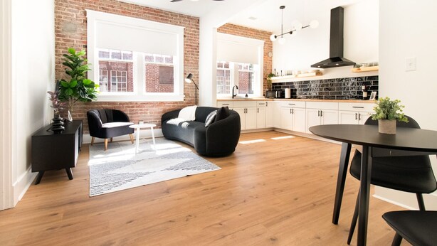A modern living room with a brick wall, large windows, a ceiling fan, and a kitchen with white cabinets and a black countertop. - Modern Loft in Lively SoMa Downtown (Little Rock)