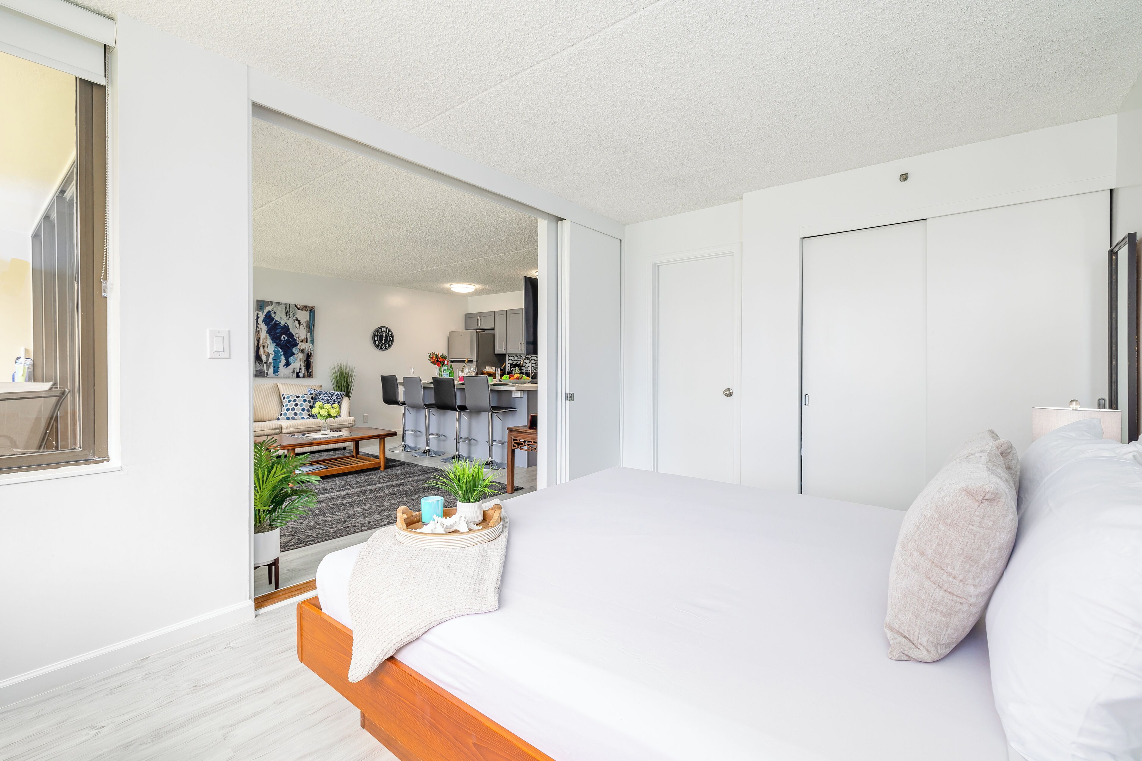 A modern bedroom with a large bed, a bedside table with a tray, and a view into the living area.