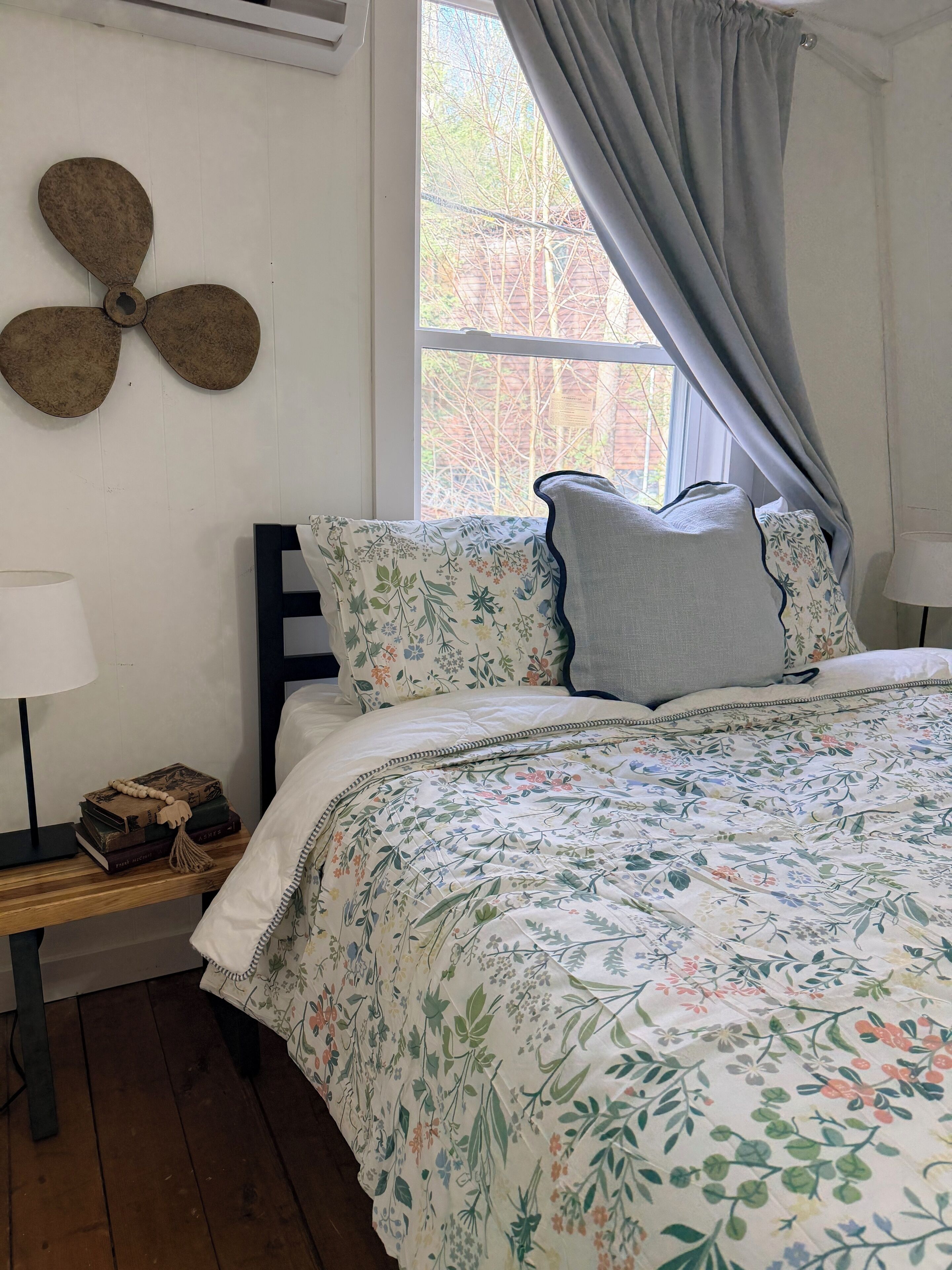 A bedroom with a bed, a bedside table with a lamp, a wall fan, and a window with curtains.