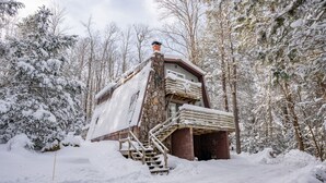A rustic cabin with a stone chimney, wooden staircase, and a snow-covered roof, surrounded by a forest. - 3-bed, charming modernized cottage tucked into wooded hillside in Deep Creek, MD (Oakland)
