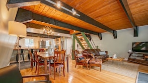 A spacious loft with a high ceiling, wooden beams, a dining area with chairs and a table, a leather sofa, a TV, and a staircase. - 3-bed, charming modernized cottage tucked into wooded hillside in Deep Creek, MD (Oakland)