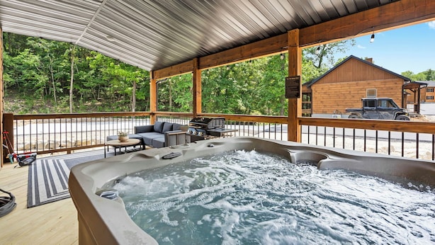 A hot tub area with a wooden deck, outdoor furniture, and a view of a forest. - Timber Valley Cabin - 4BR, Waterview, Private Hot Tub, Resort Style Pool (Hollister)