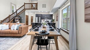 A spacious living area with a wooden dining table set for six, a brown sofa, and a staircase leading to an upper level. - Timber Valley Cabin - 4BR, Waterview, Private Hot Tub, Resort Style Pool (Hollister)