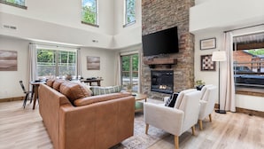 A spacious living room with a fireplace, a sofa, and a TV mounted on the wall. - Timber Valley Cabin - 4BR, Waterview, Private Hot Tub, Resort Style Pool (Hollister)
