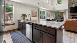 A spacious kitchen with dark cabinetry, granite countertops, and a large island. There are multiple windows providing natural light and views of the outdoors. - Timber Valley Cabin - 4BR, Waterview, Private Hot Tub, Resort Style Pool (Hollister)