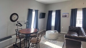 A living room with a round table, chairs, a sofa, a patterned ottoman, a wall clock, and a flat-screen TV. - Remodeled downtown studio with remarkable views! (Luverne)