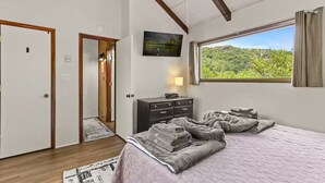 A bedroom with a bed, a dresser, a nightstand, a window with a view of greenery, and a door leading to another room.