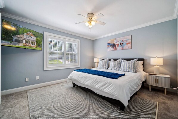 A bedroom with a bed, a ceiling fan, a window with blinds, a painting, and a nightstand with a lamp. - Lakefront Home with Private Pool and Dock- Great for Families! (Mooresville)