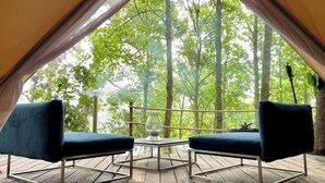 Ein Zelt mit zwei dunkelblauen Stühlen und einem kleinen Tisch, auf einer Holzterrasse mit Blick auf einen Wald.