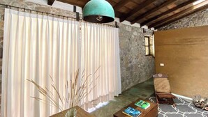 Une pièce rustique avec un mur en pierre, des poutres en bois au plafond, un luminaire suspendu, une chaise en bois, une petite table avec des livres et une plante en pot.