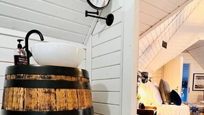 A rustic interior with a wooden barrel, a white sink, a mirror, a wicker basket, and a candle on the floor. - Stunning Tiny House w/ Outdoor Tub & Views (Pontsticill)