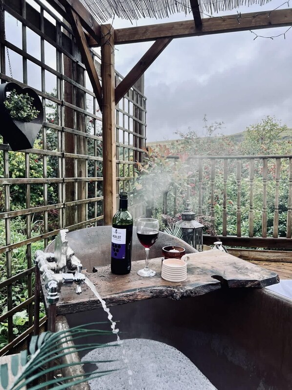 Een jacuzzi met een fles Merlot wijn en glazen, omringd door een houten pergola met uitzicht op groen.