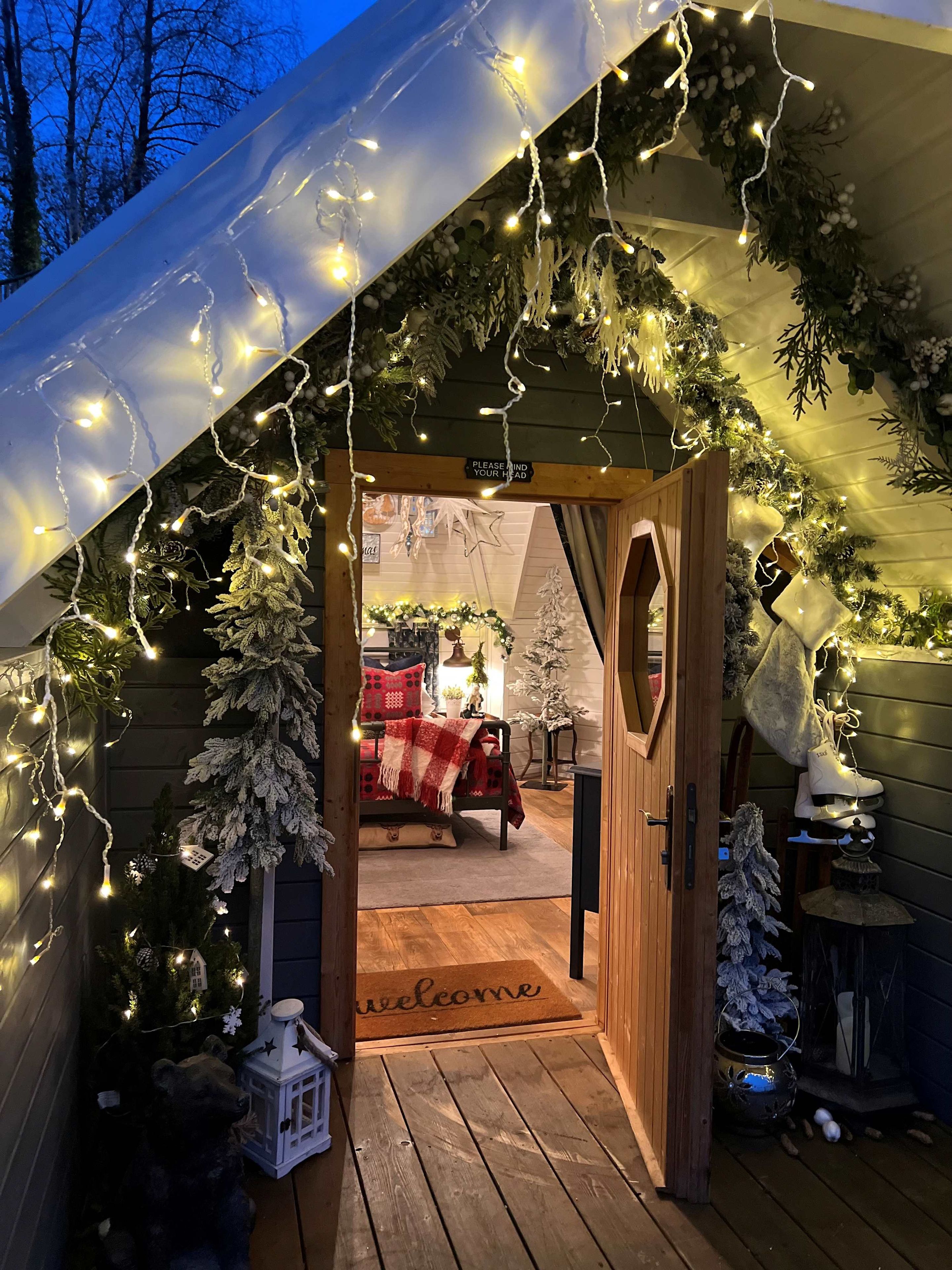 Entree van een houten hutje, met een bord waarop 'Zoek je sleutel graag hier', een welkomstmat en een kerstboom.