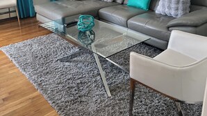 Ein modernes Wohnzimmer mit einem grauen Sofa, einem Glastisch, einem grauen Hochflor Teppich und einem weißen Sessel.