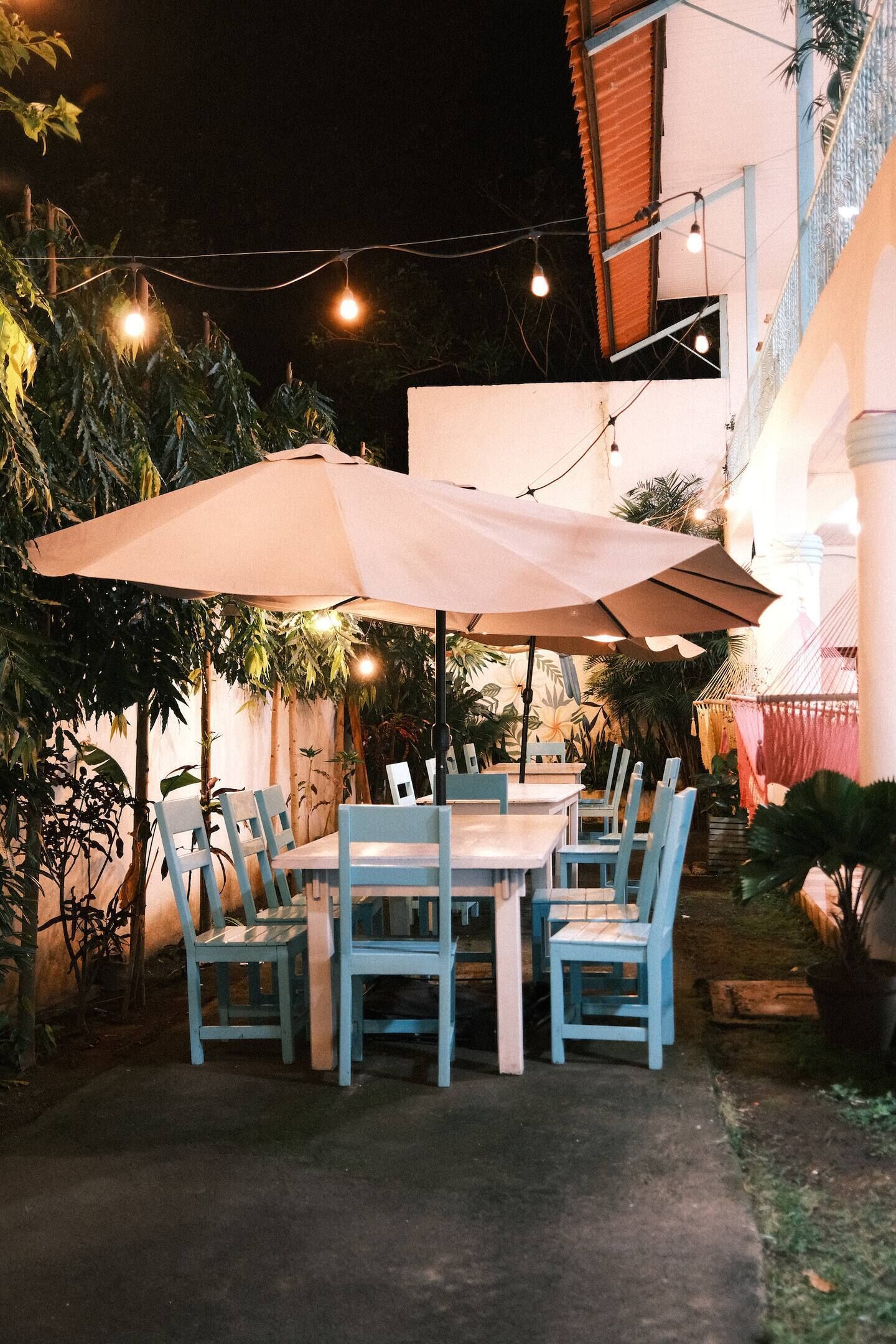 An outdoor dining area with a table and chairs under a large umbrella, surrounded by potted plants and string lights.