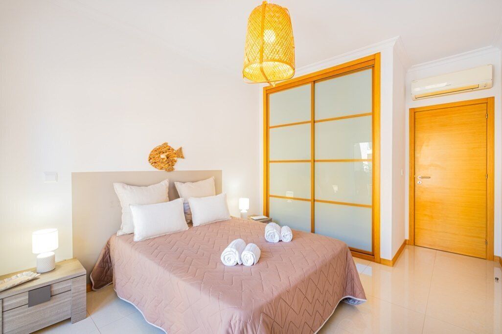 Una habitación de hotel con cama, mesita de noche, lámpara, ventana grande con persianas y puerta de madera.