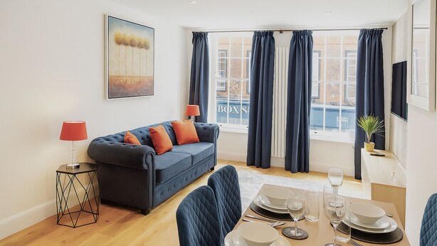 Una sala de estar moderna con un sofá, una mesa de comedor y sillas. Hay una ventana con cortinas, un cuadro en la pared y una lámpara sobre una mesita.