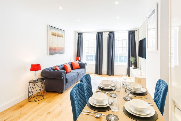 A modern living room with a sofa, a dining table set for four, and a TV mounted on the wall. - 
Elegant Windsor Flat -Short Walk to Windsor Castle, River Thames &Train Station (Windsor)