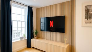 Una sala de estar moderna con un televisor de pantalla plana montado en una pared de madera, una ventana con cortinas, un banco de madera y un cuadro enmarcado en la pared.