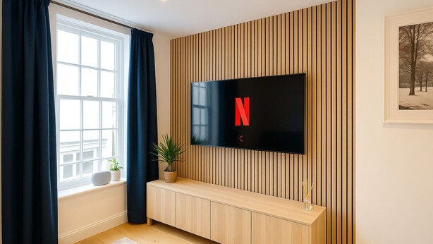 Een moderne woonkamer met een flatscreen televisie aan een houten wand, een raam met gordijnen, een houten bankje en een ingelijst beeld aan de muur.