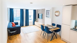 Een moderne woonkamer met een bank, een eettafel voor vier personen en een keuken met een koelkast en kastjes.