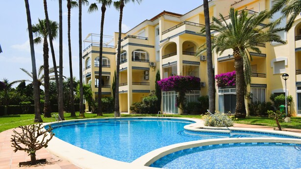 A swimming pool surrounded by palm trees and a yellow building with balconies. - Anacasa Royal Playa les Marines AP1213 (Denia)