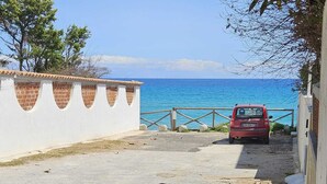 A coastal road with a red car parked by the sea, a white wall with decorative cutouts, and a fence leading to the beach.