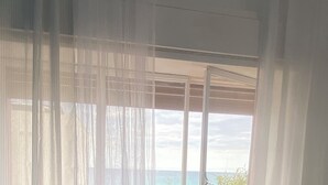 A room with a large window covered by sheer curtains, a bed with a textured white mattress, and a view of the sea and greenery outside.