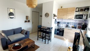Ein modernes Wohnzimmer mit einer Couch, einem Couchtisch und einem Essbereich mit Stühlen. Man sieht eine Küche mit Holzausstattung und einer Mikrowelle.