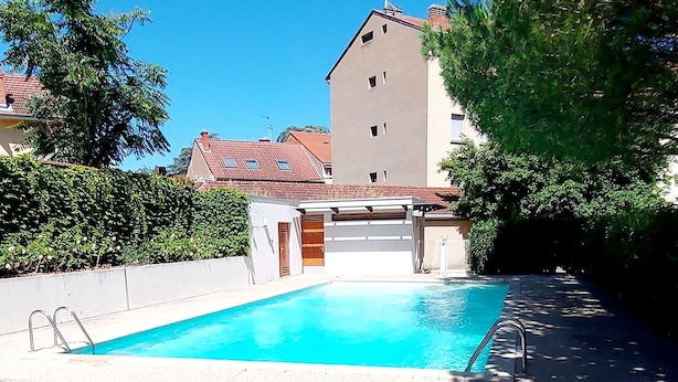 Ein Swimmingpool mit klarem, blauem Wasser, umgeben von einer weißen Mauer und einer Holzterrasse. Im Hintergrund ein mehrstöckiges Gebäude mit rotem Dach und schlichter Fassade.
