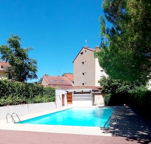 Una piscina con agua cristalina azul, rodeada por un muro blanco y una terraza de madera. En el fondo, se aprecia un edificio de varios pisos con techo rojo y fachada plana.