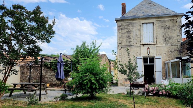 A rustic stone house with a tiled roof, a wooden bench, and a garden with flowering plants.