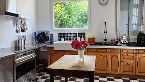 A kitchen with a checkered floor, a window with a view of greenery, a clock on the wall, and a vase with flowers on the table.