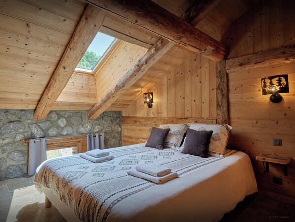A wooden cabin bedroom with a sloped ceiling, stone wall, and a bed with patterned bedding. - Chalet Marijulo - Stylish property with hot tub and terrace - OVO Network (La Giettaz)