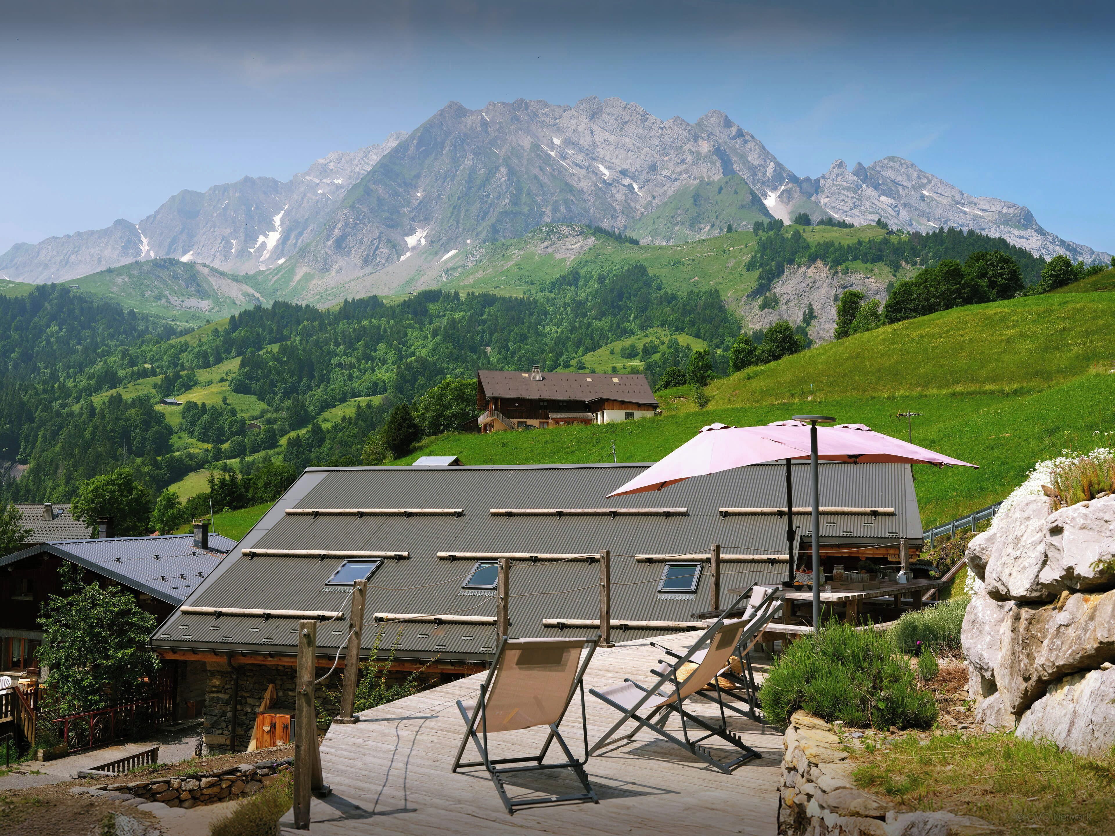 A mountain lodge with a deck and lounge chairs, overlooking a valley and mountains.