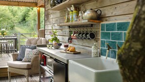 A kitchen with wooden shelves, hanging pots, a dining area with wicker furniture, and a view of greenery outside. - The Hut Rawsons in Levens (Kendal)