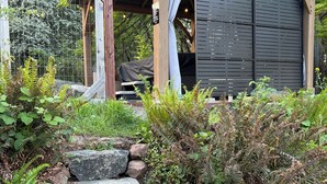 En rustik träkonstruktion med ett takförsett sittområde, omgiven av en naturlig trädgård med stentrappor som leder upp till den.