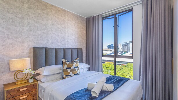 Ein Hotelzimmer mit einem großen Bett, einem Nachttisch, einer Lampe und Blick auf eine Stadtlandschaft durch ein großes Fenster.