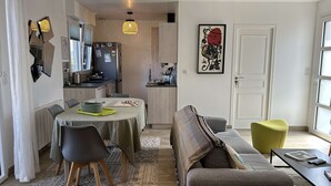 Un salon moderne comprenant un canapé, des fauteuils, une table basse et un coin repas avec une table et des chaises. Il y a également un coin cuisine avec un réfrigérateur et un évier. Un tableau est accroché au mur, et une porte avec une fenêtre est visible.