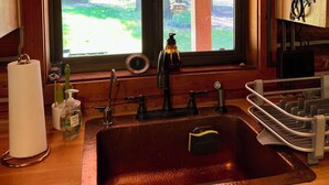Interior de una cabaña rústica de madera con un ventanal grande, un lavamanos de cobre y techo de madera.