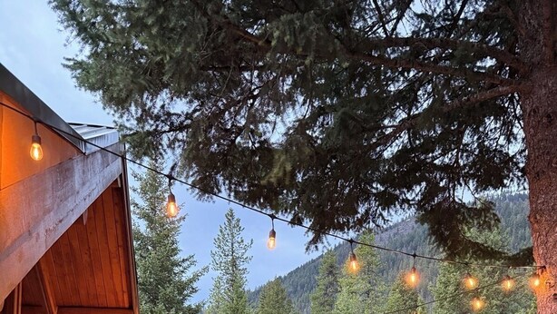 A wooden cabin with a deck, string lights, a hot tub, and a forest backdrop.
