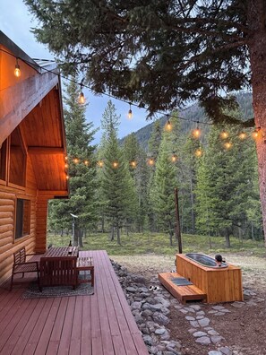 Une cabane en bois dotée d’une terrasse, de guirlandes lumineuses, d’un jacuzzi et surplombant une forêt.