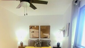 Un dormitorio con cama, ventilador de techo, dos cuadros en la pared, mesita de noche con lámpara y ventana con cortinas.