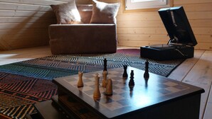 Une pièce chaleureuse avec un plafond en bois, une fenêtre, un canapé avec des coussins, un jeu d’échecs et un tourne-disque.