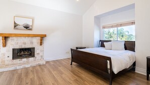 Ein Schlafzimmer mit Kamin aus Stein, einem Bett mit Holz Kopfteil, einem Fenster mit Blick auf Grünflächen und einem gerahmten Bild an der Wand.