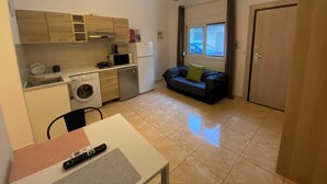 Eine kleine Küche mit Waschmaschine, Mikrowelle und kleinem Kühlschrank. Ein Sofa mit Kissen und ein Fenster mit Vorhängen.