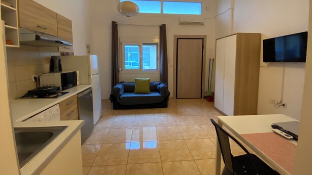 Eine kompakte Küche mit Mikrowelle, Herd und Schränken, eine Essecke mit Tisch und Stühlen, ein Sofa mit grünem Kissen, ein an der Wand befestigtes Fernsehgerät und ein großes Fenster mit Vorhängen.