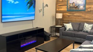 A modern living room with a flat-screen TV, a black entertainment unit with a built-in mini fireplace, a grey sofa, and a wooden accent wall with a framed painting. - New Listing!  Oasis Bungalow (Rockport)