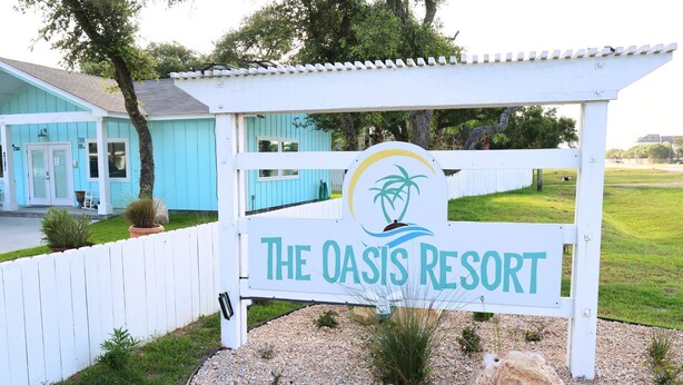 A sign for The Oasis Resort with a palm tree logo, surrounded by a white picket fence and gravel. - New Listing!  Oasis Bungalow (Rockport)
